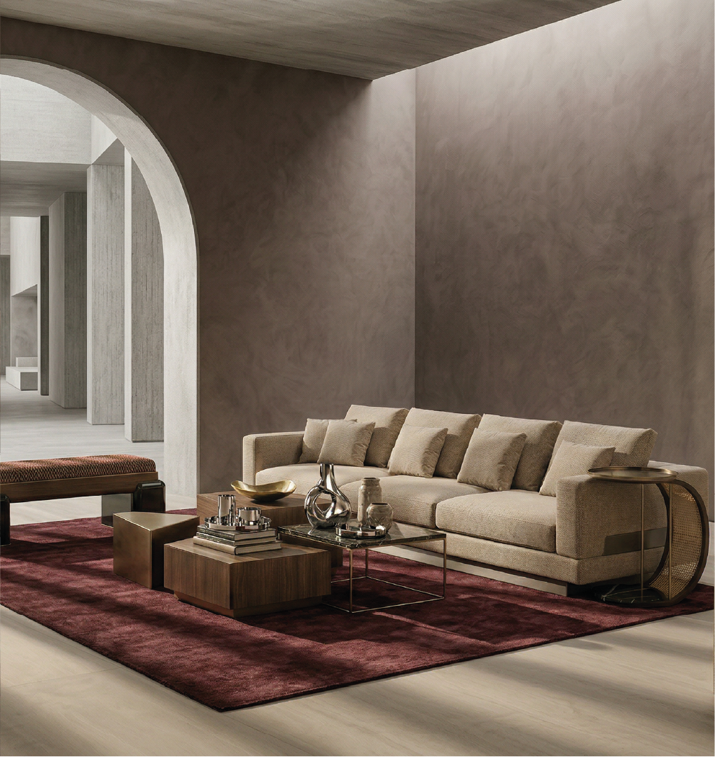 Britten Sofa in a modern, minimalistic living room with beige tones, featuring plush cushions, a dark maroon rug, and stylish metallic decor.